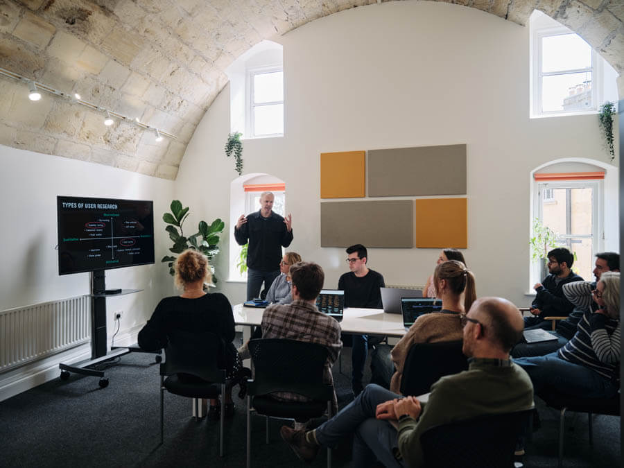 Levelling up with the UX Design Institute: How a Senior Designer filled critical knowledge gaps after years in the industry 2 Mark presenting a slide on types of user research to a small group seated around a table with laptops in a bright, modern classroom.